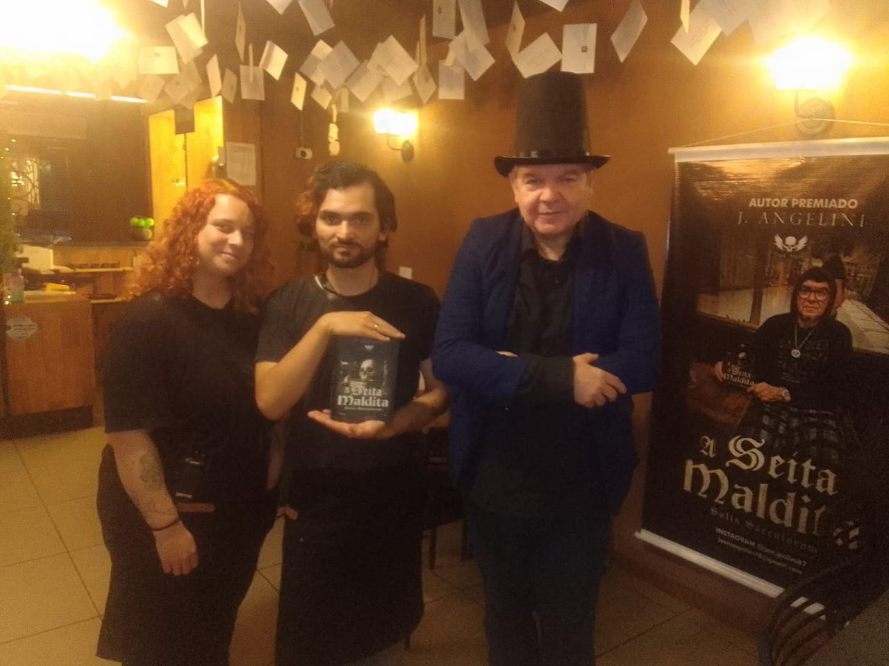 J. Angelini de cartola com casal e o banner de A Seita Maldita no lançamento