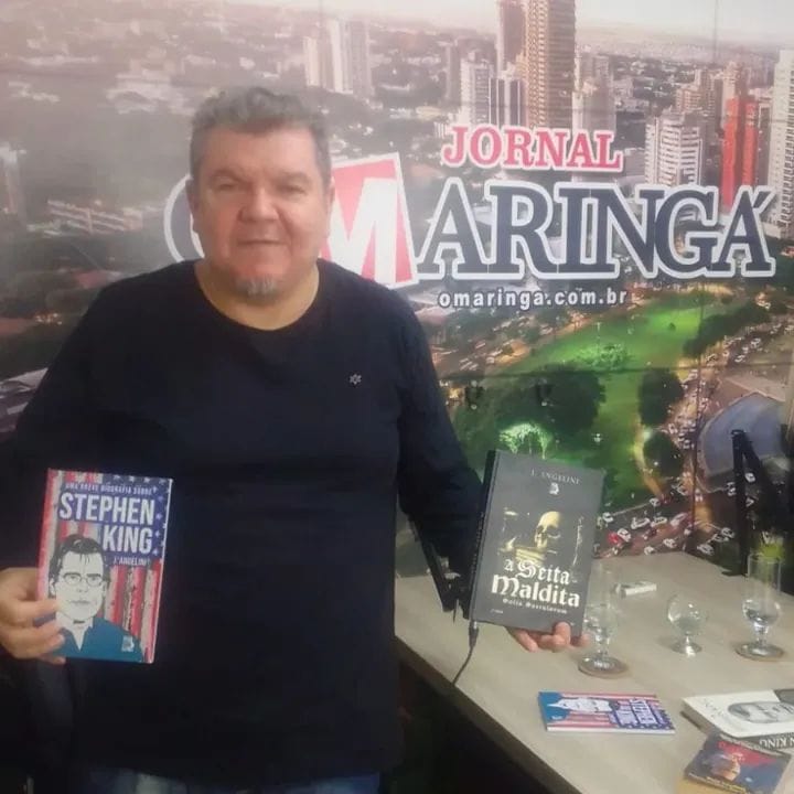 autor segurando dois livros em frente ao painel do Jornal Maringá