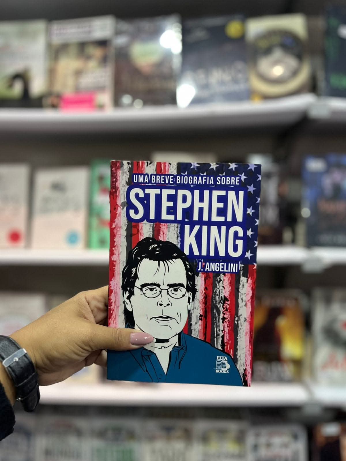 mão segurando o livro Uma breve biografia sobre Stephen King em frente à estante de livros